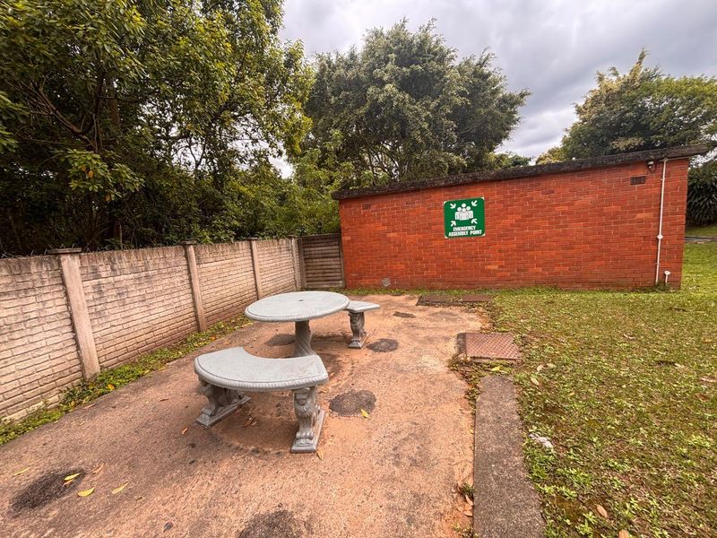 A rear outdoor area featuring a concrete paved patio, brick boundary wall, and a brick building facade with an emergency assembly point sign.