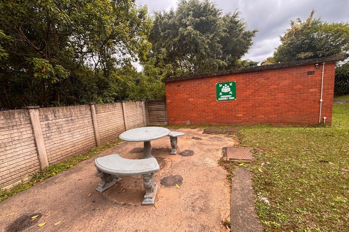 A rear outdoor area featuring a concrete paved patio, brick boundary wall, and a brick building facade with an emergency assembly point sign.
