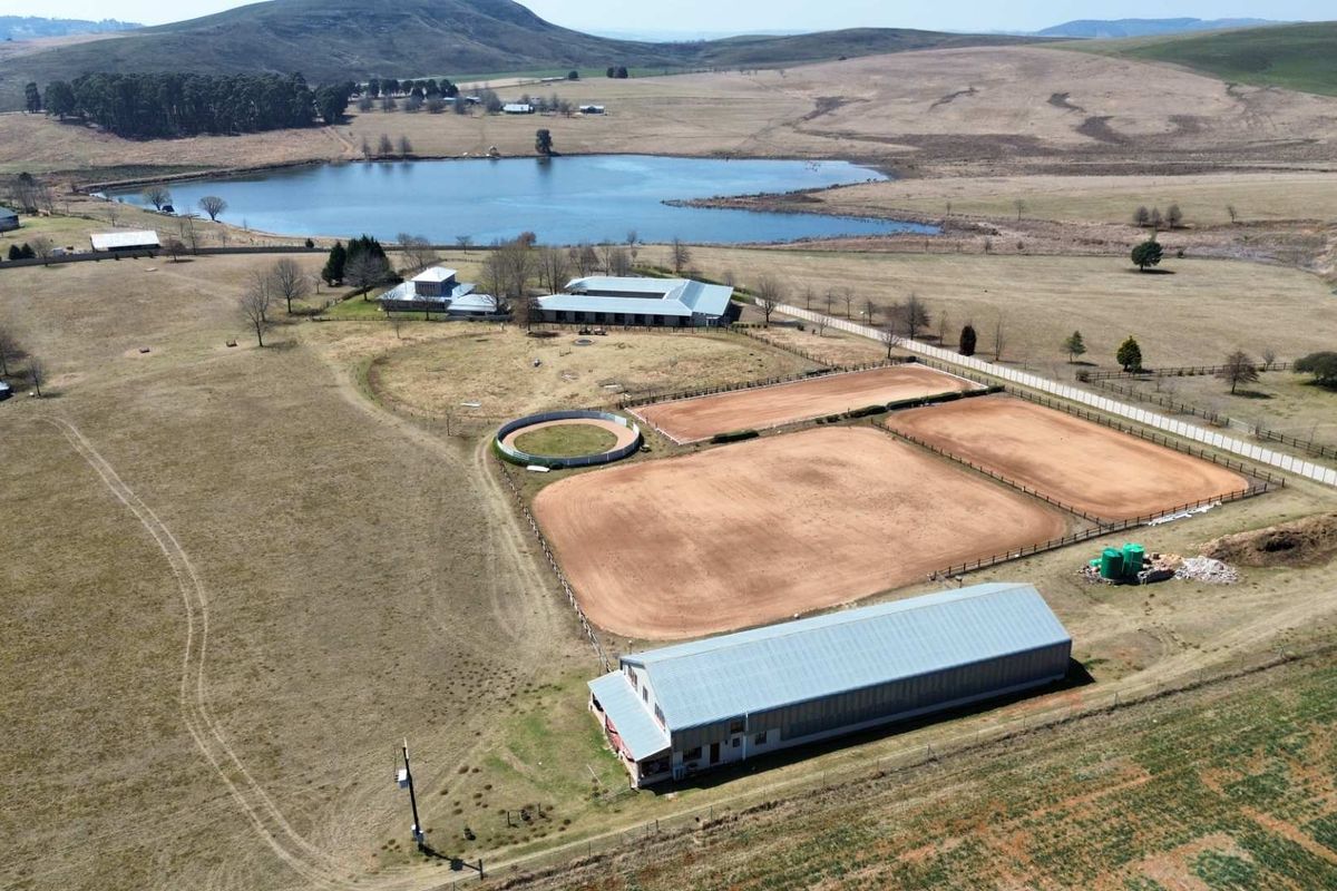 Aerial view of a large farm property featuring multiple equestrian arenas, a lunging ring, a large barn, and scenic landscape with a dam.
