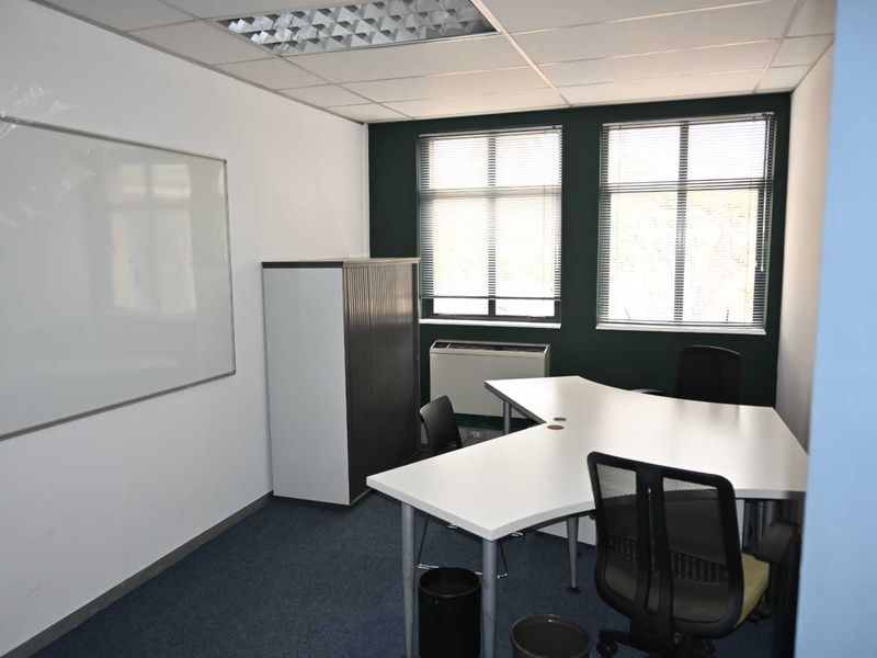 A functional office space featuring a suspended ceiling, recessed lighting, wall-mounted whiteboard, and large windows with venetian blinds.