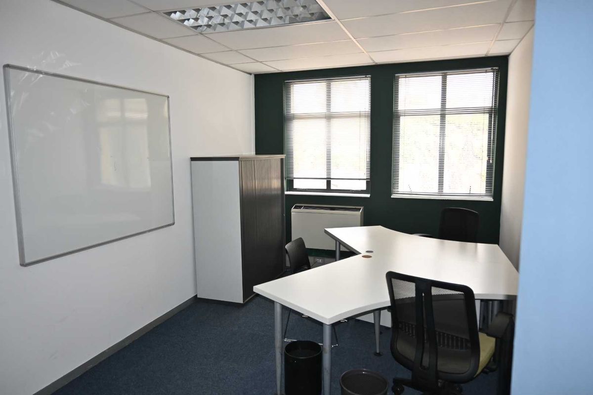 A functional office space featuring a suspended ceiling, recessed lighting, wall-mounted whiteboard, and large windows with venetian blinds.