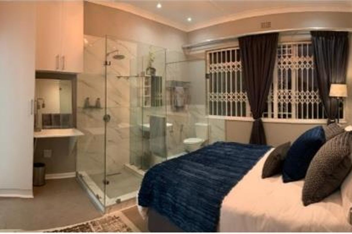 A modern bedroom featuring an integrated glass-enclosed shower, built-in vanity, and ample cupboard space, complemented by secure window treatments.
