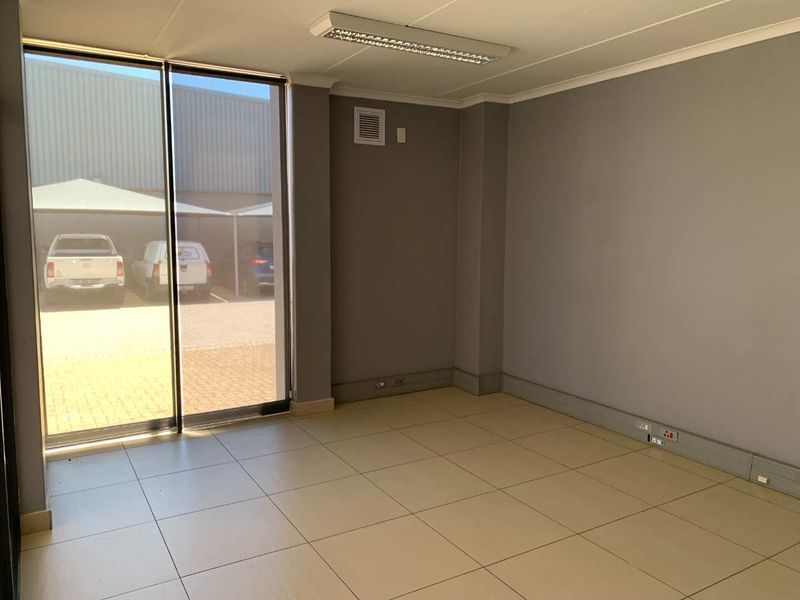 A clean, empty office space featuring neutral grey walls, ceramic tiled flooring, and a large sliding glass door providing natural light and exterior access.