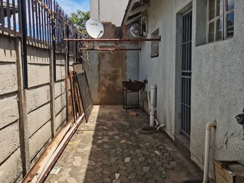 A narrow rear exterior walkway featuring paved flooring, security fencing, and external plumbing fixtures, showing signs of significant wear and maintenance requirements.