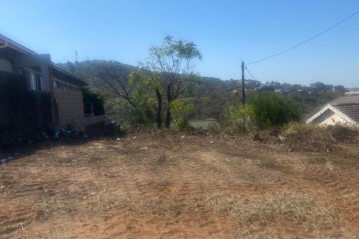 A vacant residential plot of land featuring cleared ground, adjacent to existing residential structures and local utility infrastructure.