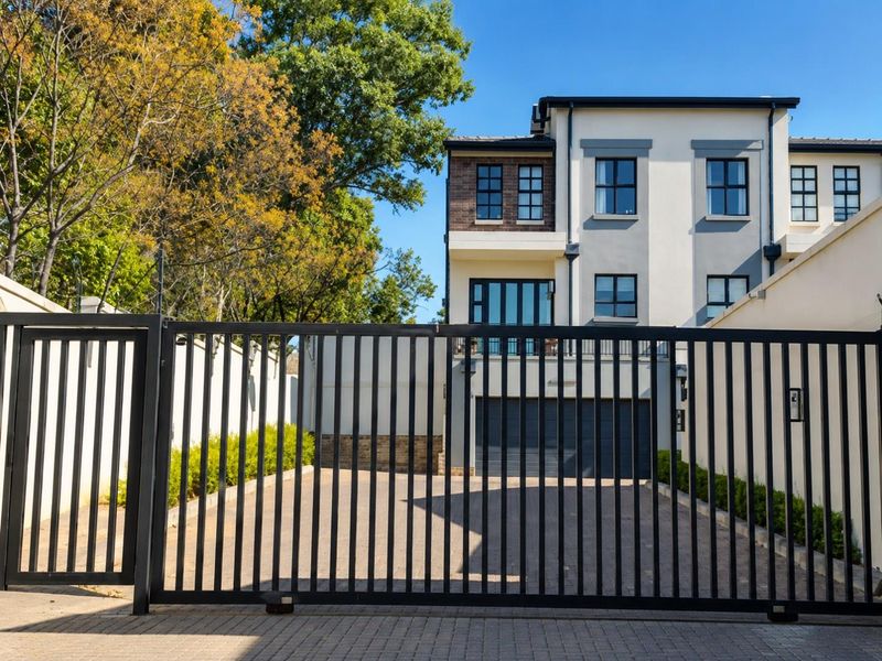 Front view of a modern multi-story townhouse featuring a secure metal gate, paved driveway, integrated double garage, and a private balcony.