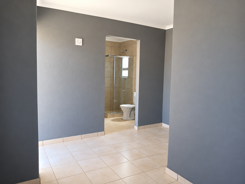 A clean, empty bedroom featuring neutral grey walls, tiled flooring, and an en-suite bathroom with a glass shower enclosure and toilet.