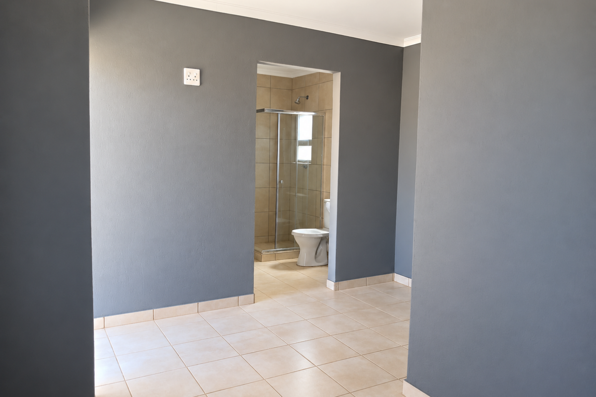 A clean, empty bedroom featuring neutral grey walls, tiled flooring, and an en-suite bathroom with a glass shower enclosure and toilet.