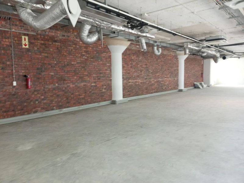 An open-plan office space featuring industrial-style exposed brick walls, concrete columns, and exposed HVAC ducting with polished concrete flooring.