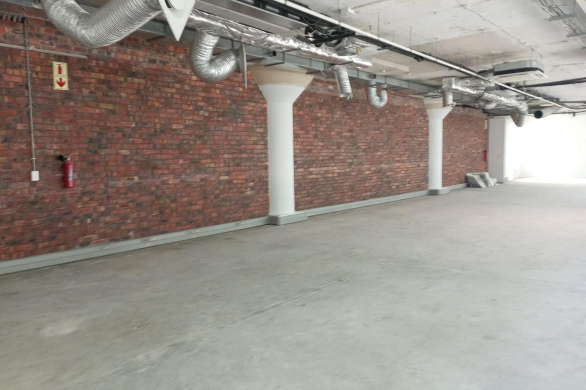 An open-plan office space featuring industrial-style exposed brick walls, concrete columns, and exposed HVAC ducting with polished concrete flooring.