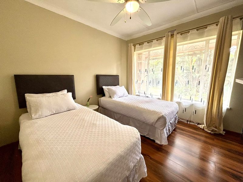 A well-lit bedroom featuring hardwood flooring, a ceiling fan, and large windows with security bars, providing a comfortable and functional living space.