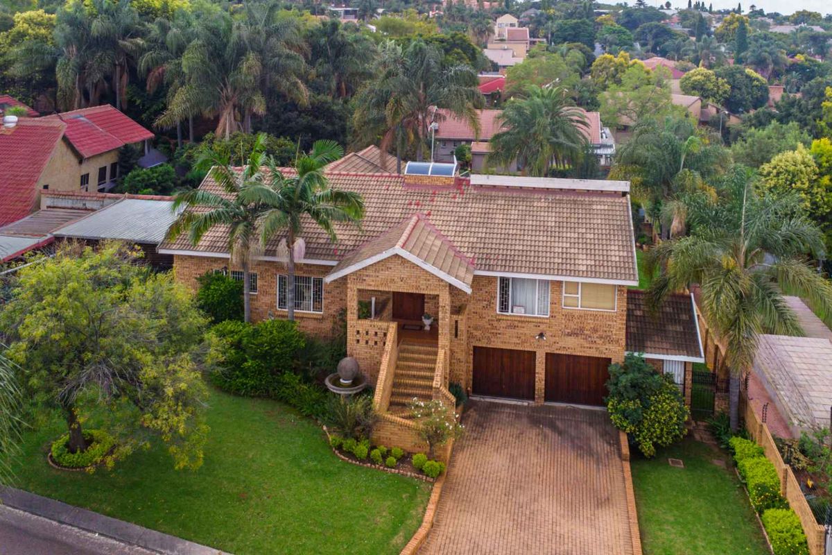 Aerial view of a brick-faced residential house featuring a tiled roof, double garage, paved driveway, and established garden in a suburban setting.