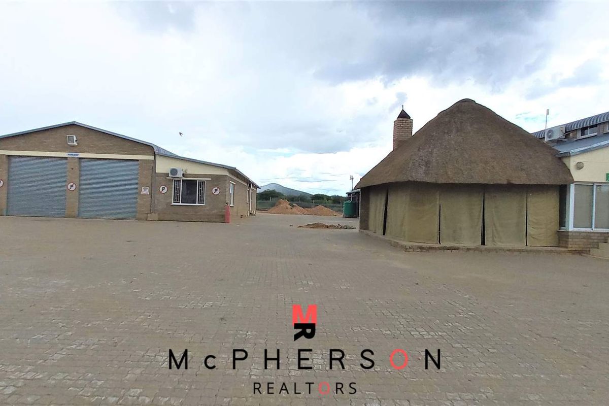 Exterior view of an industrial property featuring a large paved yard, a workshop building with roller shutter doors, and a secondary structure with thatch roofing.