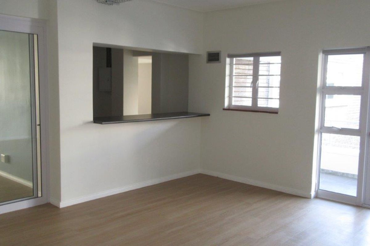 An empty, well-lit living area featuring modern laminate flooring, a convenient serving hatch to the kitchen, and access to an outdoor space via sliding doors.