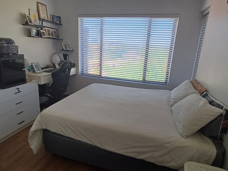 A functional bedroom featuring laminate flooring and large windows fitted with venetian blinds, offering natural light and exterior views.