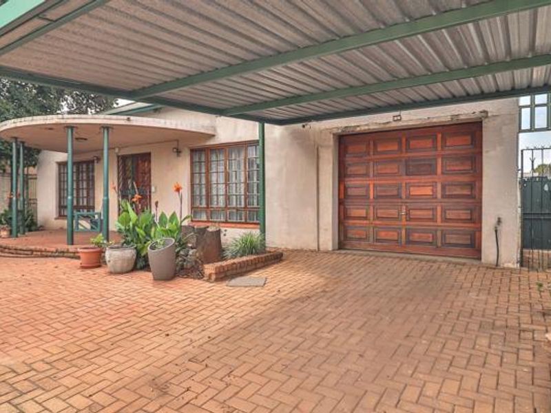 Front exterior view featuring a paved driveway, covered carport, wooden garage door, and a covered patio area with established garden beds.