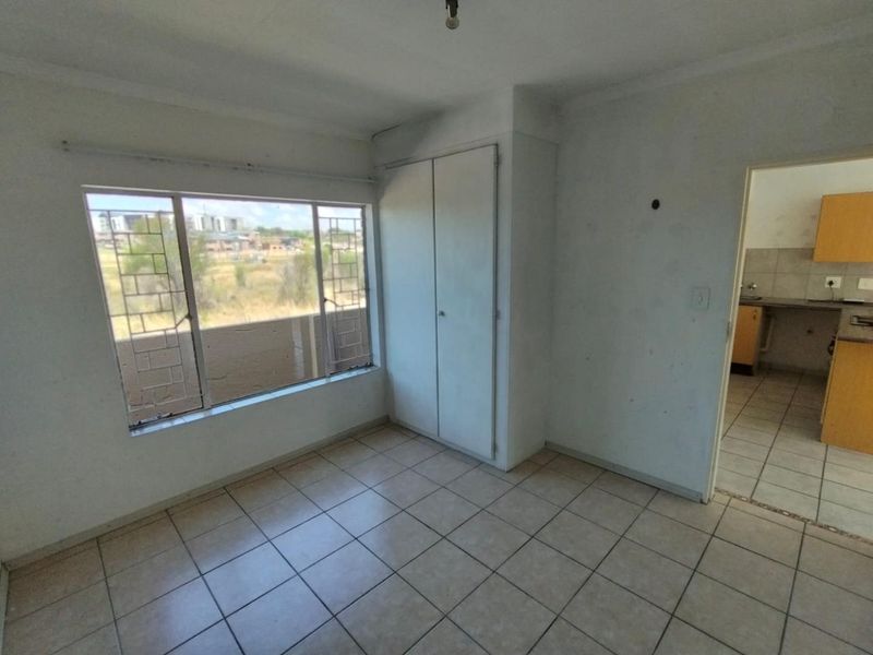 An empty bedroom featuring tiled flooring, a built-in wardrobe, and security bars on the window, with a view into the adjacent kitchen area.