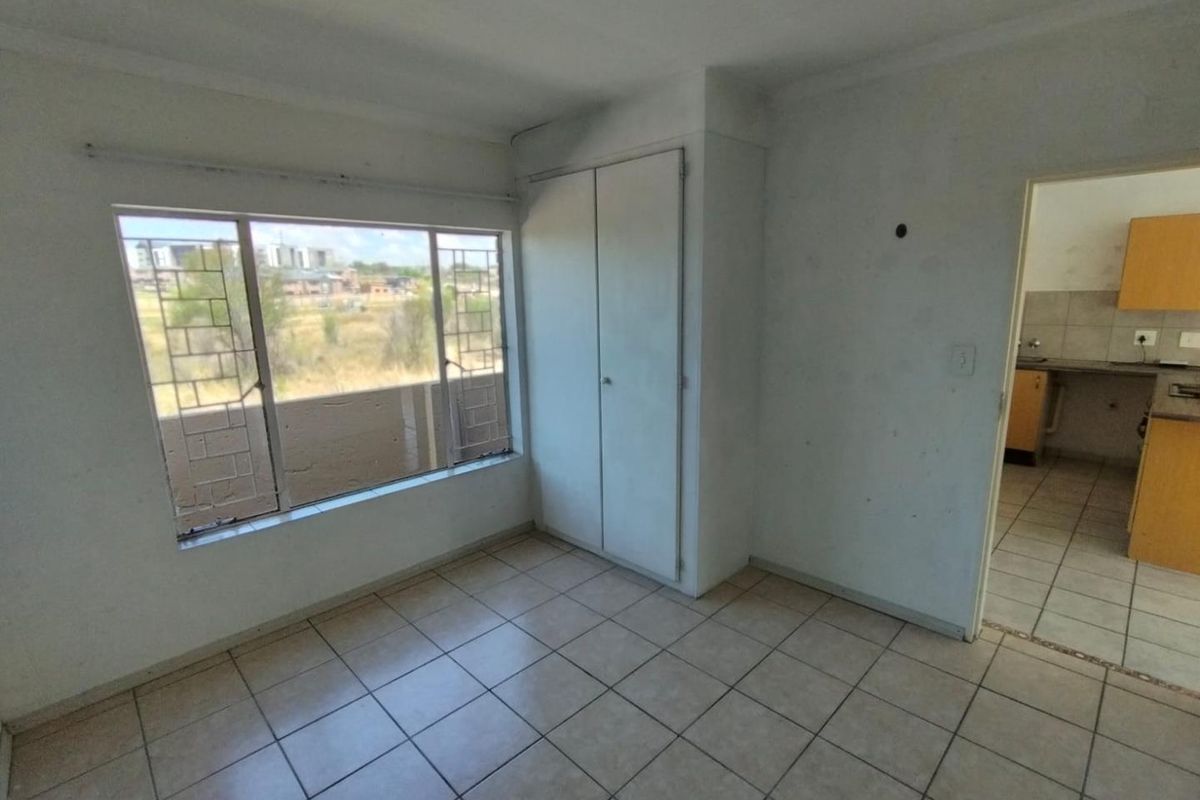 An empty bedroom featuring tiled flooring, a built-in wardrobe, and security bars on the window, with a view into the adjacent kitchen area.