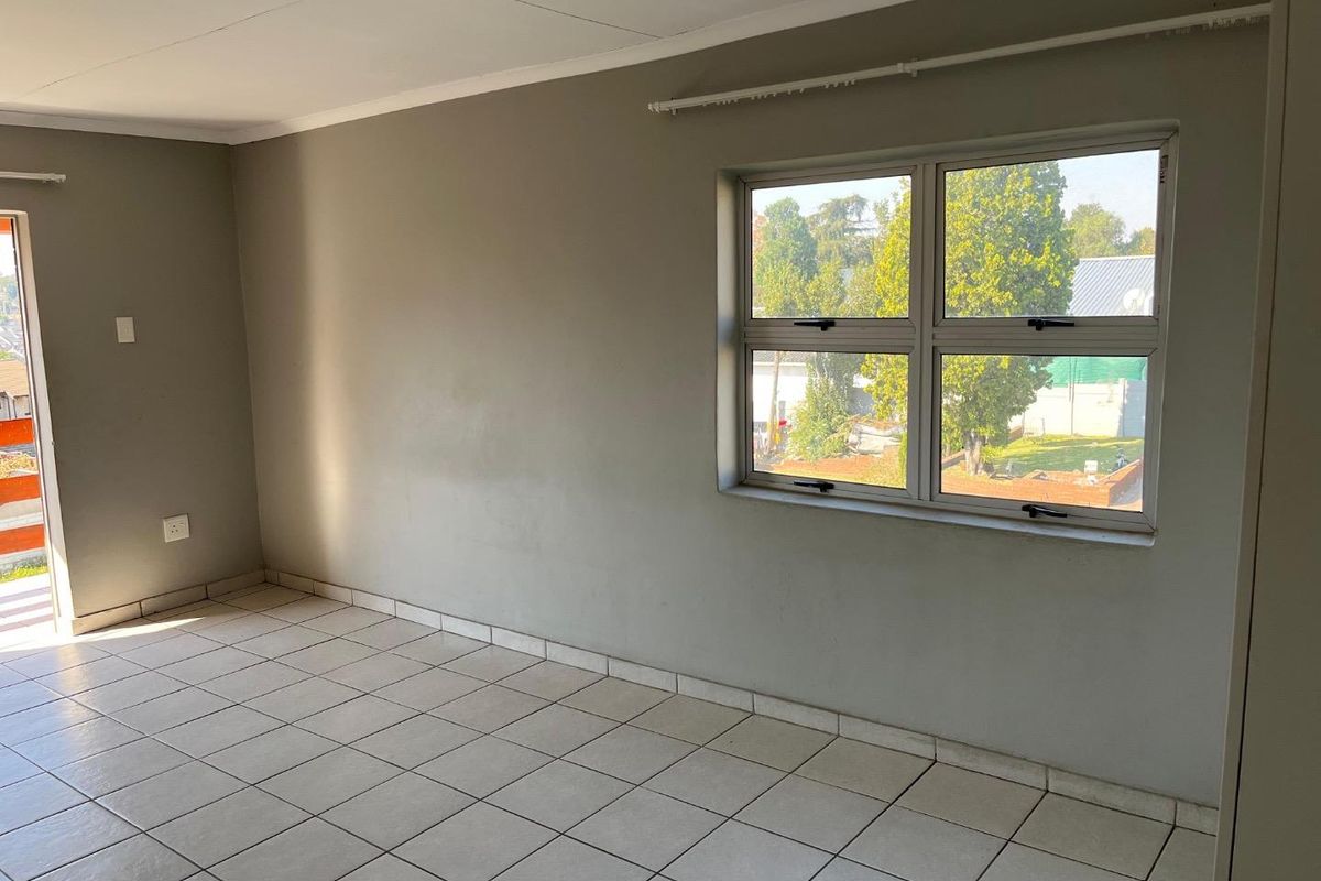 A bright, empty bedroom featuring neutral grey walls, tiled flooring, and large windows providing natural light and garden views.