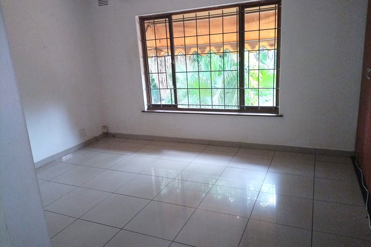 A bright, empty bedroom featuring tiled flooring, built-in cupboards, and a large window fitted with security bars and an external awning.