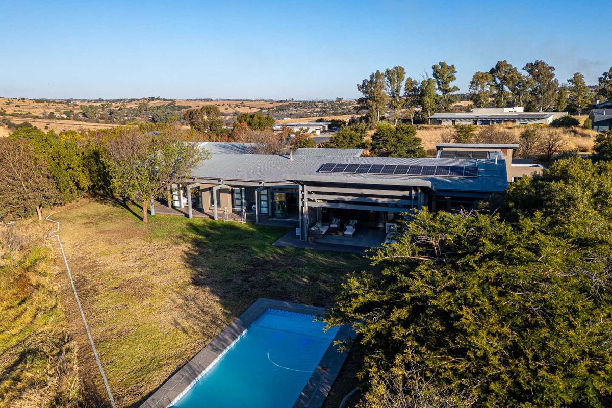Aerial view of a modern residence featuring a swimming pool, expansive lawn, and solar panels installed on the metal roof.