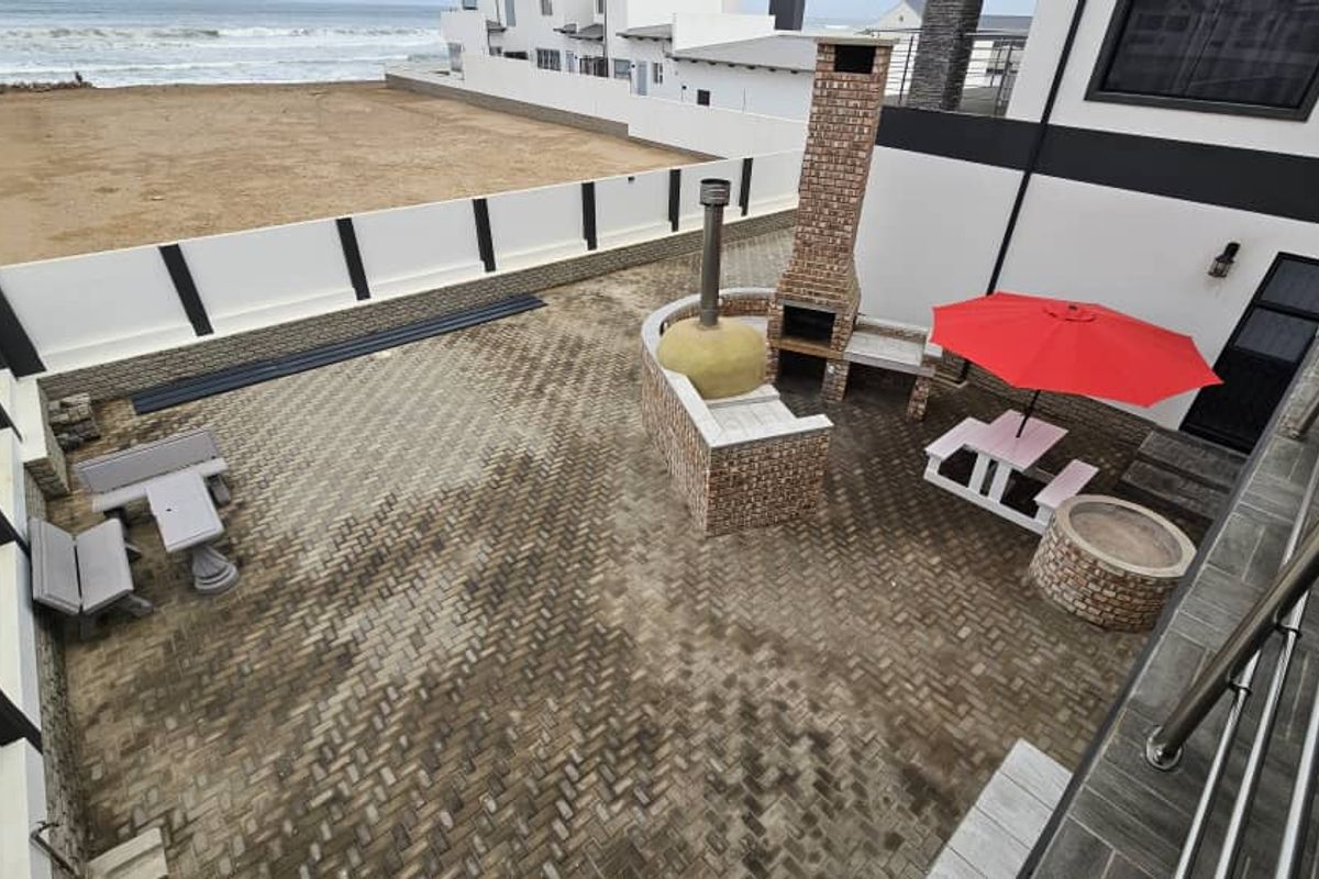 An elevated view of a paved rear courtyard featuring a built-in brick braai, pizza oven, and fire pit area overlooking the ocean.