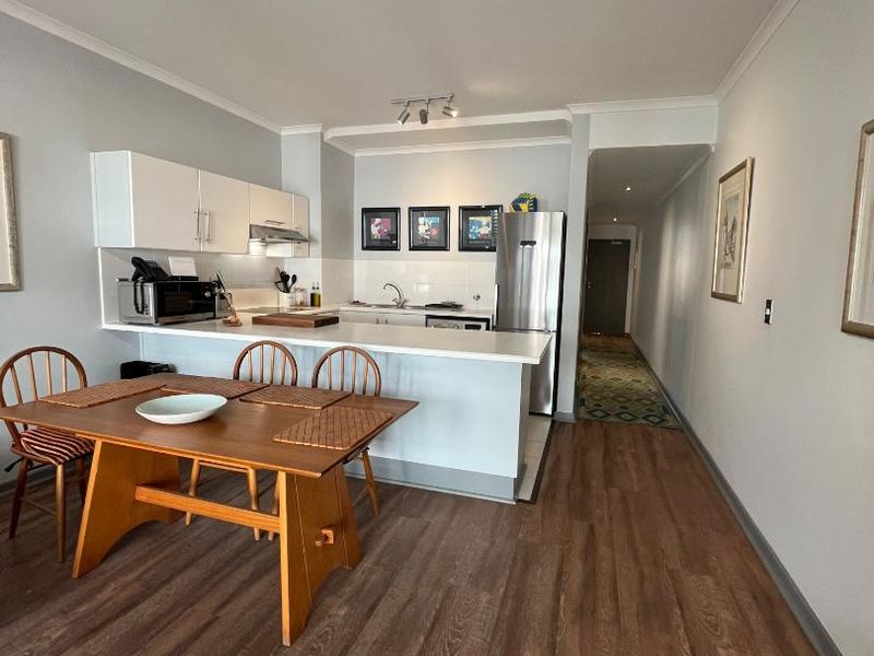 Open-plan kitchen and dining area featuring modern white cabinetry, a breakfast bar, and durable laminate flooring, leading into a hallway.