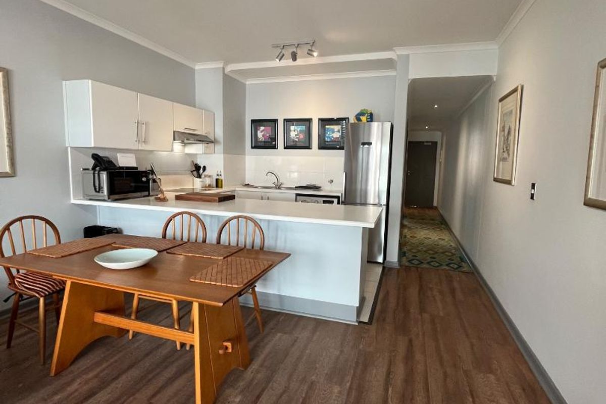 Open-plan kitchen and dining area featuring modern white cabinetry, a breakfast bar, and durable laminate flooring, leading into a hallway.