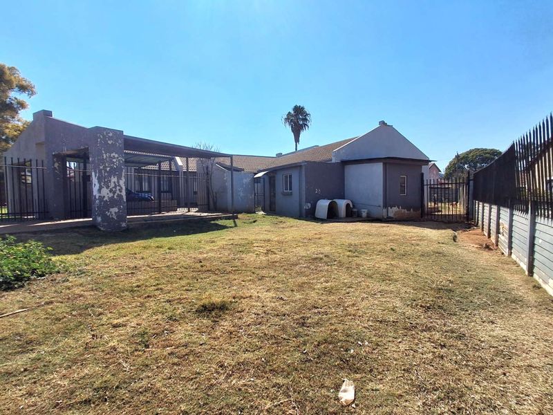 Rear view of the property featuring a large garden, covered patio area, and perimeter metal fencing with visible exterior wall maintenance required.