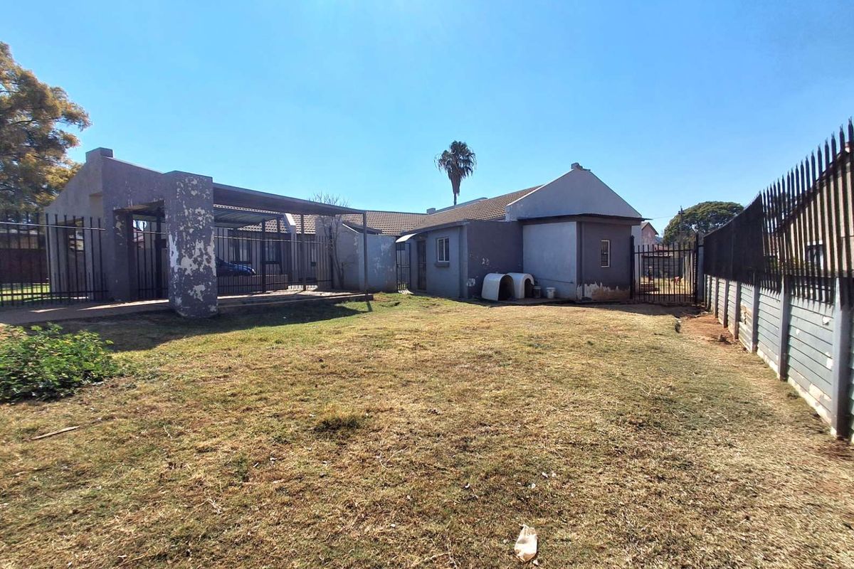 Rear view of the property featuring a large garden, covered patio area, and perimeter metal fencing with visible exterior wall maintenance required.