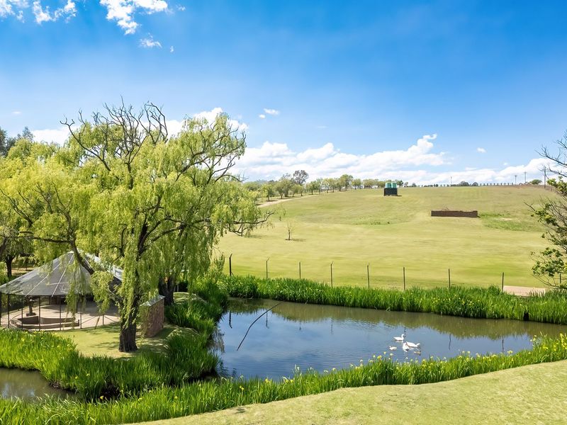 A scenic view of a landscaped pond, mature willow trees, and a gazebo set against a backdrop of expansive green hills.