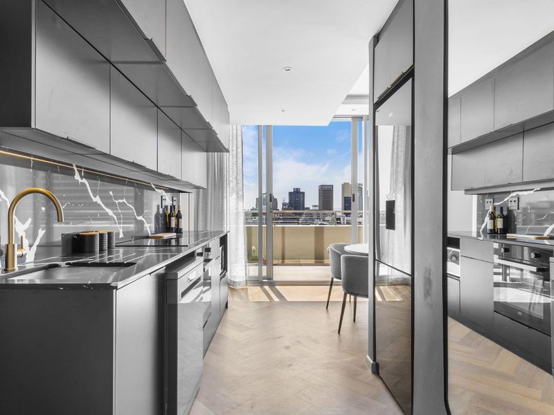 Modern galley-style kitchen featuring sleek dark cabinetry, marble-look splashback, integrated appliances, and herringbone timber flooring, opening onto a balcony with city views.
