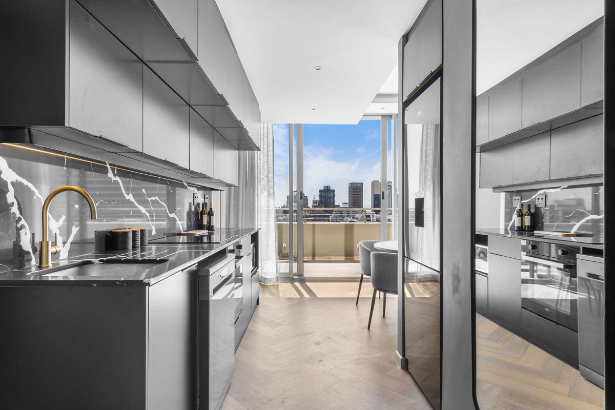 Modern galley-style kitchen featuring sleek dark cabinetry, marble-look splashback, integrated appliances, and herringbone timber flooring, opening onto a balcony with city views.