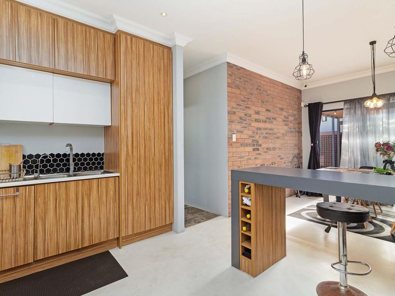Modern open-plan kitchen and dining area featuring wood-grain cabinetry, a striking exposed brick wall, hexagonal tile backsplash, and contemporary pendant lighting.