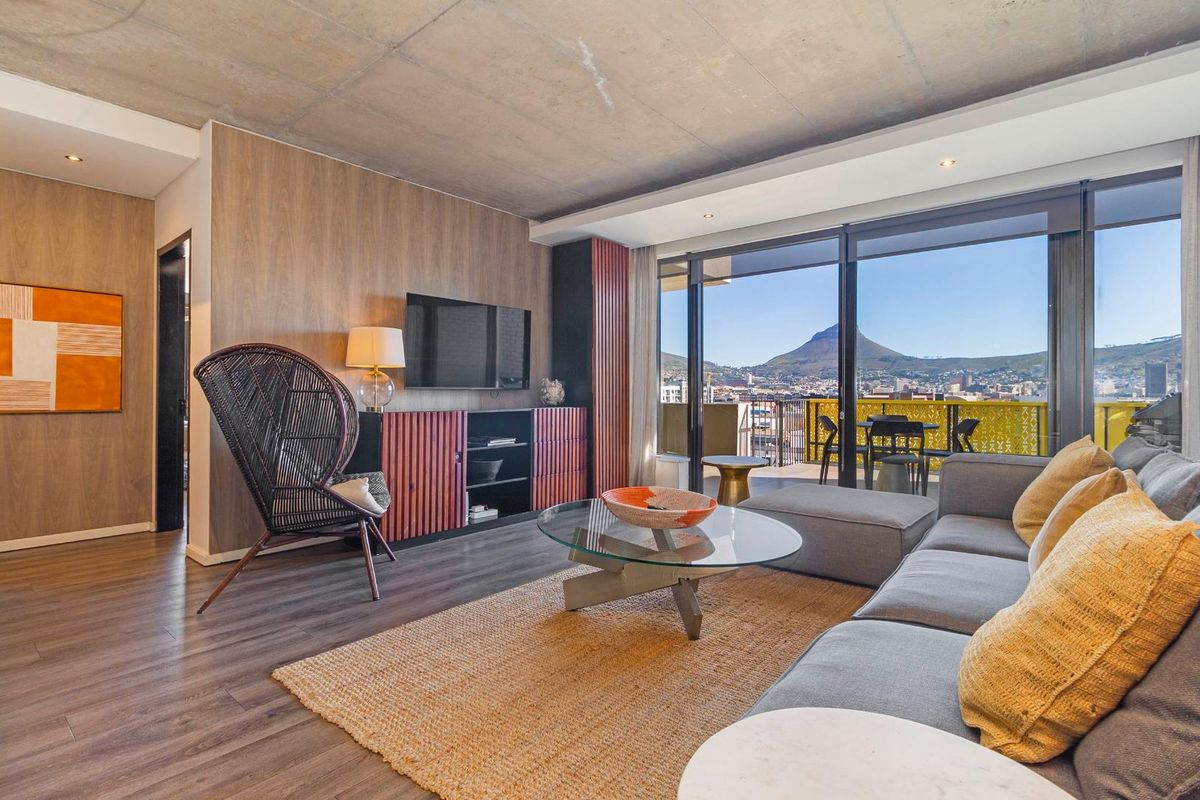 Modern living room featuring exposed concrete ceiling, wood-panelled feature wall, and large sliding glass doors opening to a balcony with mountain views.