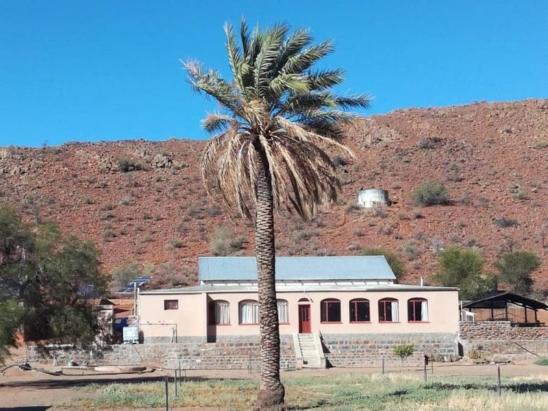 A farmstead featuring a single-story dwelling with a metal roof, stone base, and solar panels, set against a rugged mountain landscape.