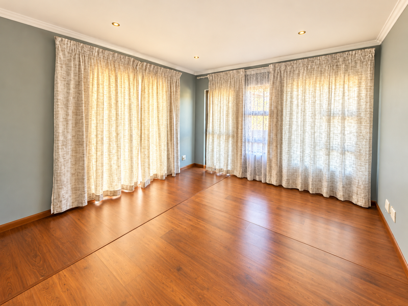 A bright and spacious empty living area featuring warm-toned laminate flooring, recessed downlights, and large windows dressed with neutral curtains.