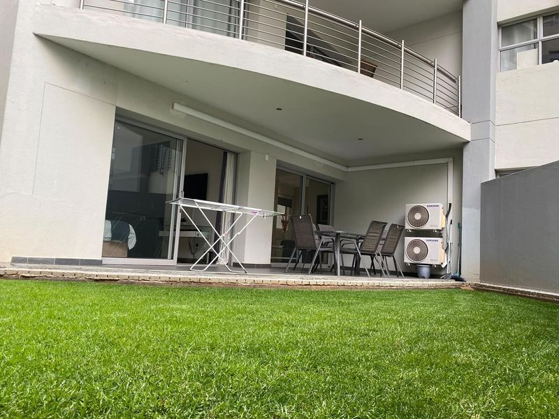 A covered ground-floor patio featuring tiled flooring, recessed lighting, and stainless steel balustrades, providing direct access to the garden area through sliding glass doors.