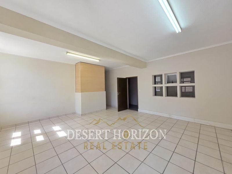 A spacious, empty open-plan industrial office or commercial area featuring neutral tiled flooring, fluorescent lighting, and an internal window partition.