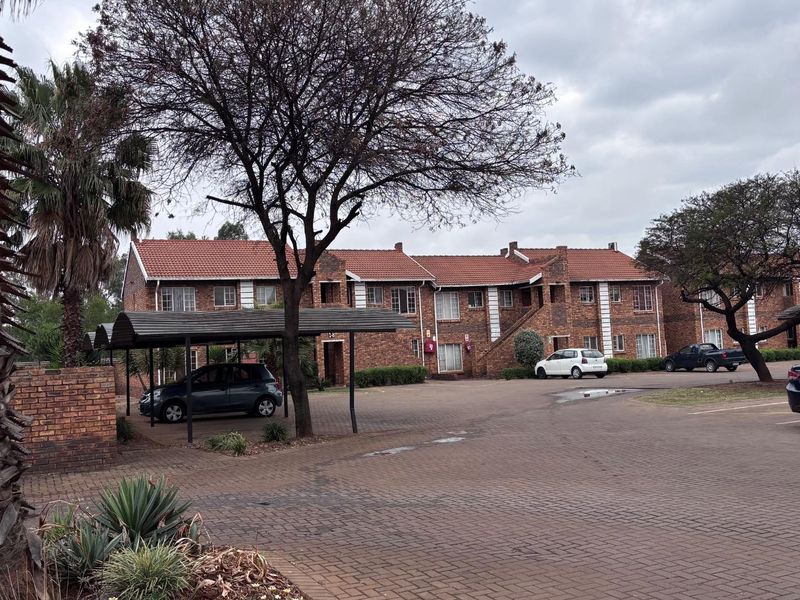 Exterior view of a brick apartment complex featuring a paved parking area, covered carports, and mature landscaping.