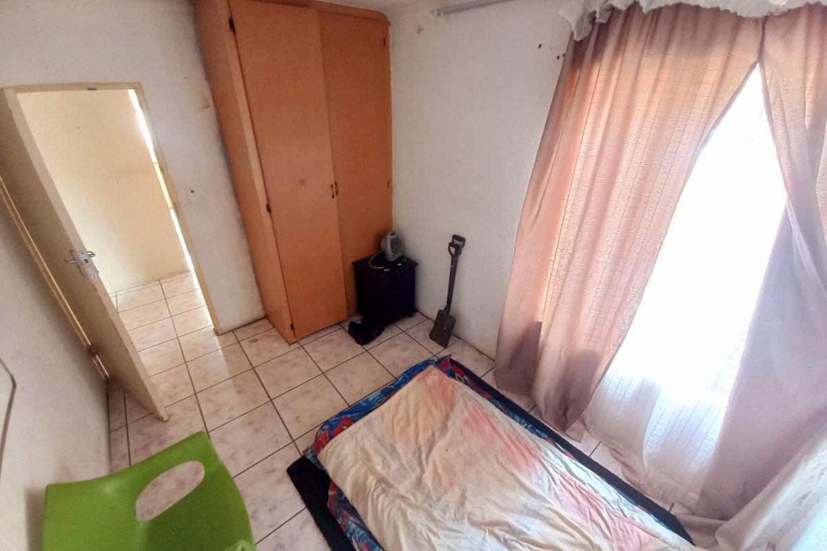 A compact bedroom featuring tiled flooring, a built-in wooden wardrobe, and a large window with curtains, requiring cosmetic updates and maintenance.