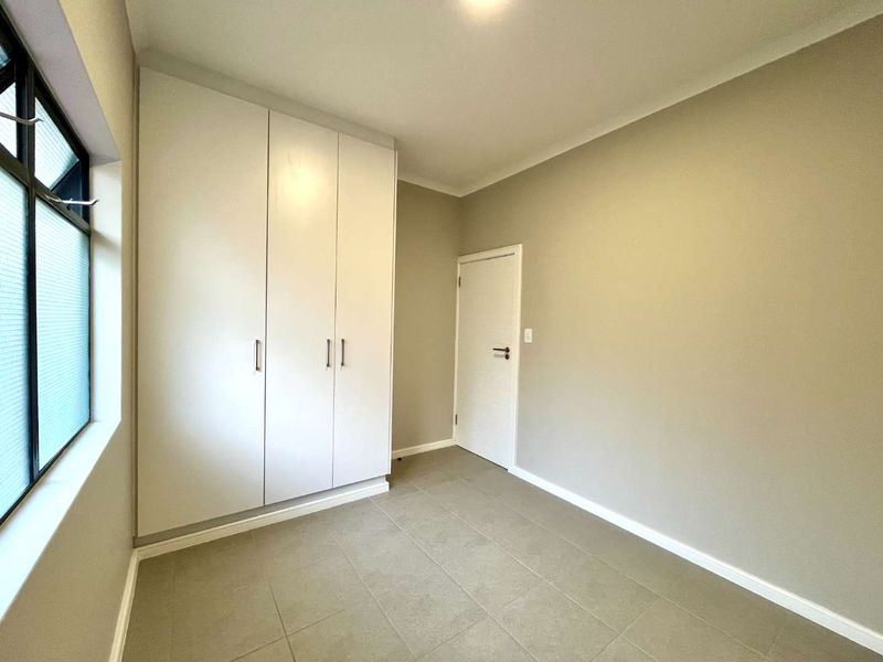A clean, neutral-toned bedroom featuring built-in cupboards, tiled flooring, and a large window, offering a bright and versatile space.
