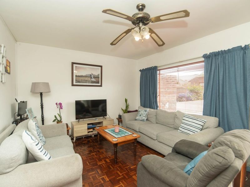 A comfortable living room featuring classic parquet flooring, a ceiling fan, and large windows with venetian blinds, offering a warm and inviting atmosphere.