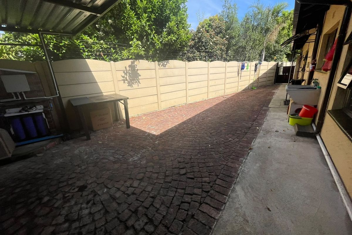 A functional rear exterior area featuring cobblestone paving, a precast concrete boundary wall, a metal carport structure, and an outdoor utility sink.