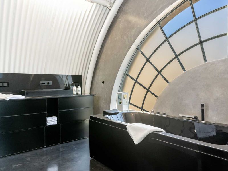 Modern bathroom featuring a sleek black built-in vanity, integrated sink, and a contemporary bathtub set against a unique arched window and corrugated metal ceiling.