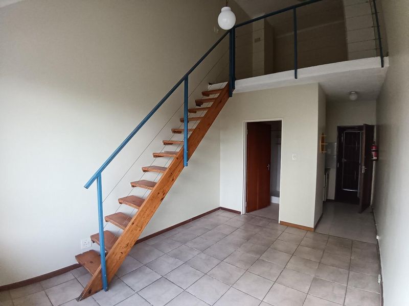 An open-plan living area featuring a timber staircase leading to a mezzanine level, finished with ceramic floor tiles and neutral wall tones.