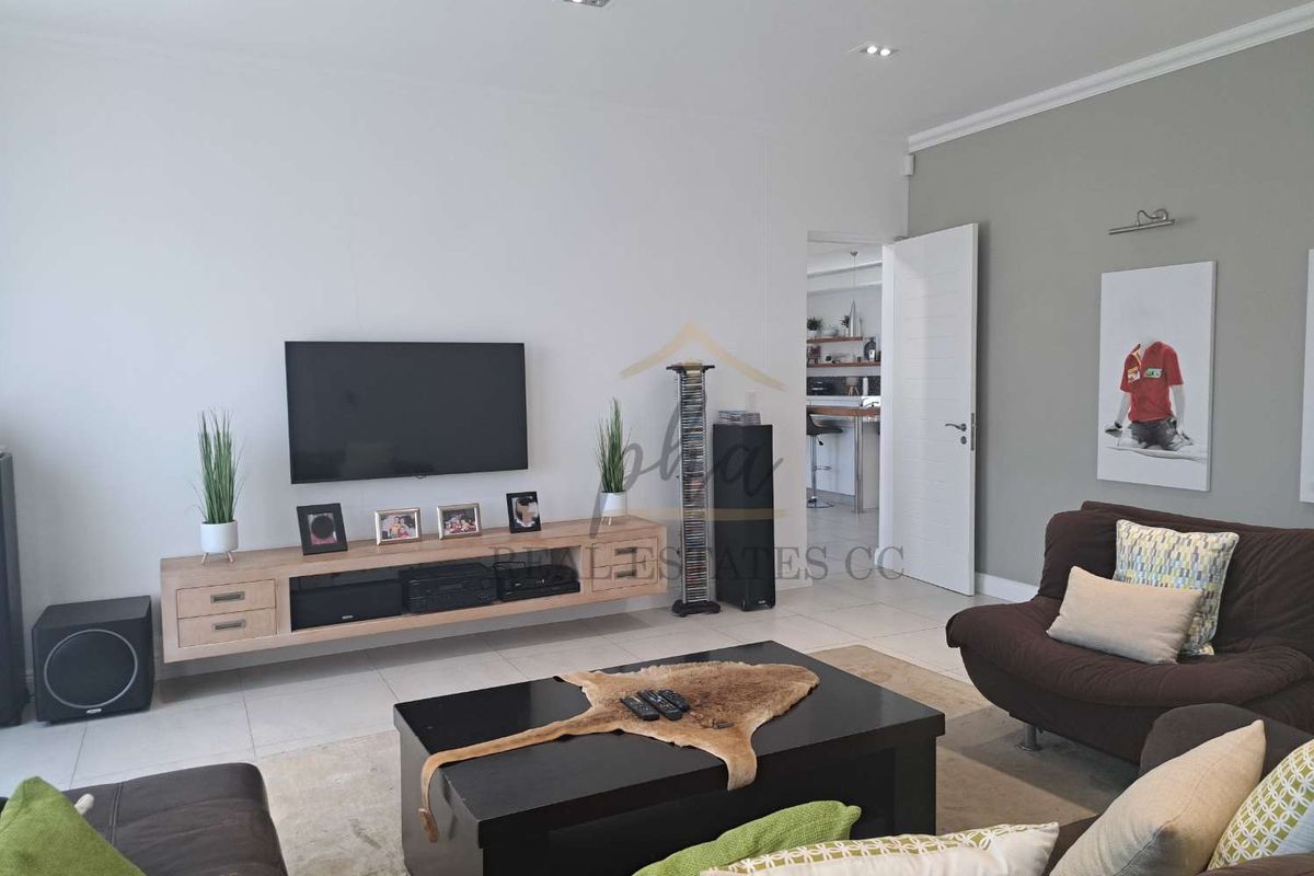 A modern living room featuring tiled flooring, recessed downlighting, and a wall-mounted media console, with a view into an adjacent kitchen area.
