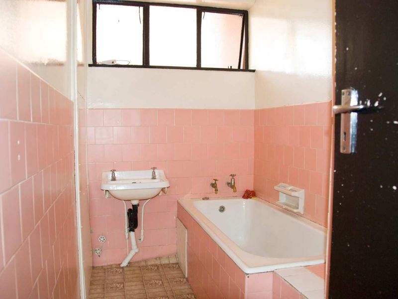 A dated bathroom featuring pink wall tiles, a built-in bathtub, and a wall-mounted basin with a high-level window.