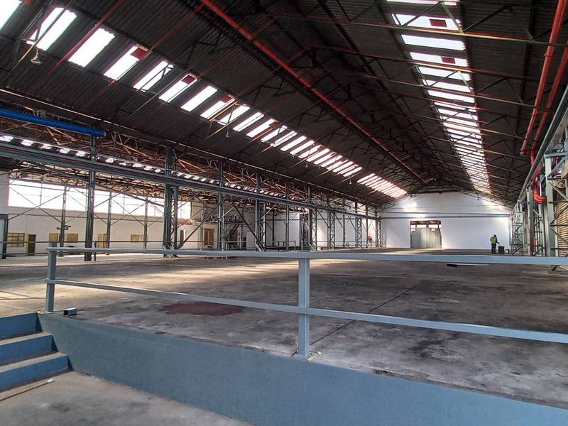 Expansive industrial warehouse space featuring high ceilings with skylights, exposed steel trusses, concrete flooring, and a raised platform area with safety railings.
