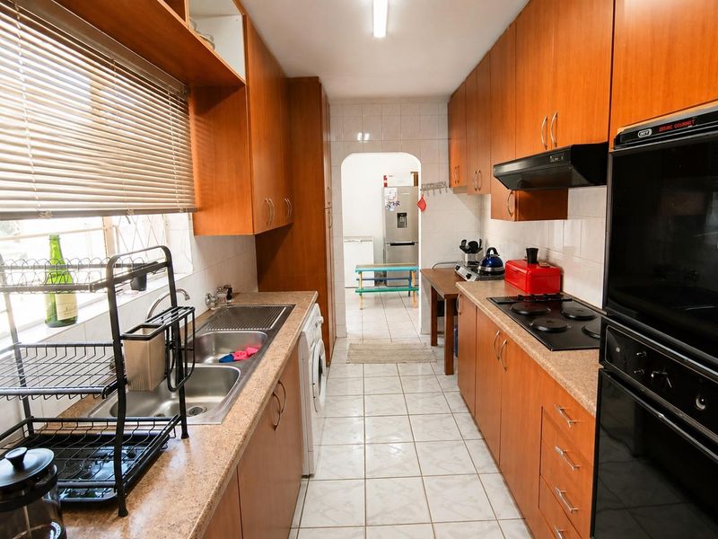 Functional galley-style kitchen featuring wood-look cabinetry, built-in electric oven and hob, double sink, and tiled flooring with direct access to a secondary utility area.